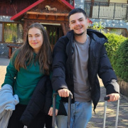 Two people stand outside a building with suitcases, smiling at the camera. They are dressed warmly, suggesting a travel or holiday setting. The building has a rustic appearance with stone and wood elements.
