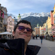 A person takes a selfie on a busy street with colourful buildings and snow-capped mountains in the background.