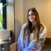 A young woman with long brown hair is sitting on a wooden chair by a window. She is wearing a blue and white striped shirt over a white top, with sunglasses resting on her head. A potted plant is on the windowsill, and the room has a warm, cosy atmosphere.