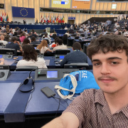 Young participant takes a selfie inside the European Parliament's hemicycle during the European Youth Event (EYE) 2025 in Strasbourg. The chamber is filled with young people from across Europe, and a screen displays the EYE 2025 logo. EU member state flags are visible in the background.