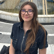 Young girl with glasses and long hair standing in front of a waterfountain.