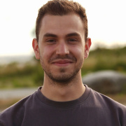 Marcos looking into the camera with a smile outdoors wearing a dark grey shirt standing outside with some green in the background