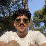 A young man with curly dark hair and a moustache wearing round green-tinted sunglasses and a white T-shirt, sitting outdoors among tall pine trees on a sunny day, looking at the camera