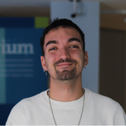 A young person with short dark hair, facial hair, and earrings is smiling warmly at the camera while wearing a white top. The background is softly blurred with blue tones.