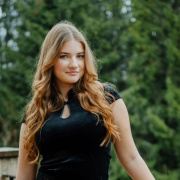 Young girl with long, wavy hair in a black outfit standing confidently on a balcony with a forest background. She has a relaxed, serene expression.