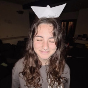 A young woman with long wavy brown hair smiles with her eyes closed while balancing a white paper boat on her head. She has a nose ring and a septum piercing and is wearing a grey sweatshirt. The photo appears to be taken indoors during a group activity or workshop, with dim lighting and chairs visible in the background. The paper boat and her relaxed expression create a playful and light-hearted atmosphere.