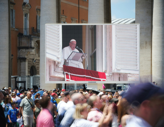 Screen displays a religious figure speaking from a balcony; crowd assembled below, with historic building and columns in the background.