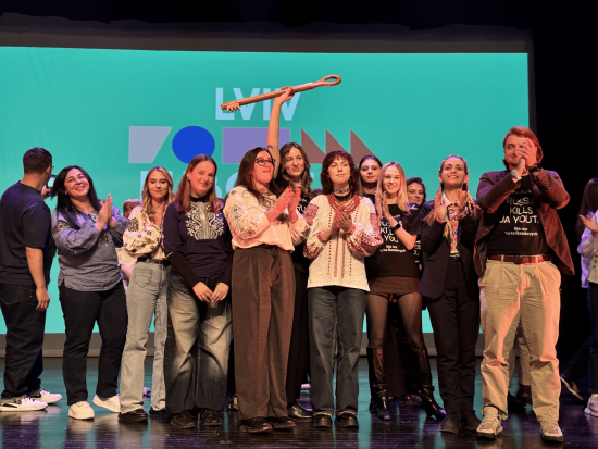 Group on stage in front of blue-green backdrop, clapping and smiling. Centre person holds large key. Some wear embroidered shirts; others have text shirts.