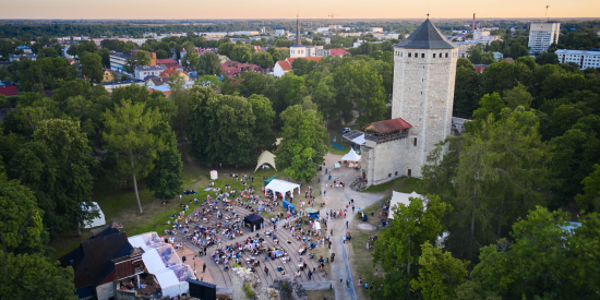 Aerial view of a public event near a stone tower surrounded by trees. People are gathered in an outdoor seating area with a backdrop of residential buildings.