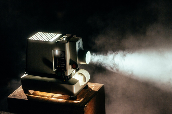 Vintage film projector on a wooden box, projecting a bright beam of light through smoky air in a dimly lit room. The side buttons contribute to its retro look.