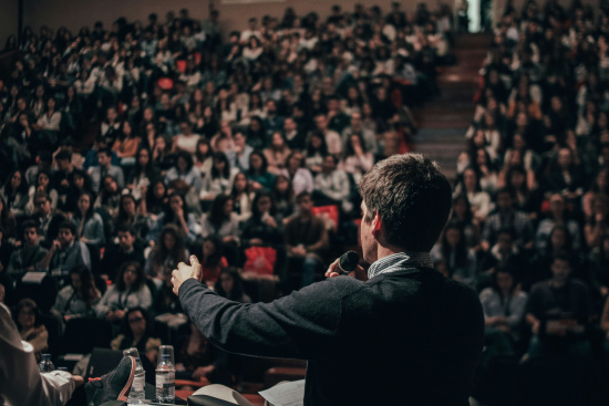 Man with a microphone speaking to a large, attentive audience in a crowded auditorium.