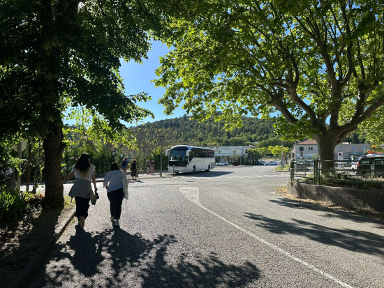 Two individuals walk under trees towards a parked white tour bus. A hotel sign and buildings are seen against a backdrop of hills.