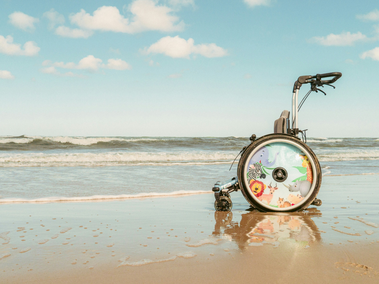 A wheelchair with animal illustrations on its wheels sits on a sandy beach, partially in the sea. The waves are gently rolling in under a blue sky with scattered clouds.
