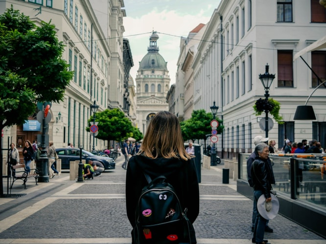 Person with a black backpack stands on a European cobblestone street facing a domed building. The street has elegant buildings and trees, with people walking and engaging in various activities.