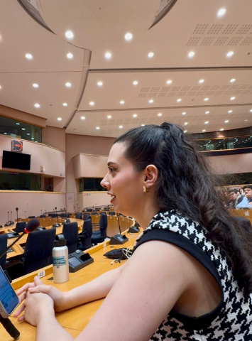 Woman with dark hair and a houndstooth top speaks into a microphone in a large conference room. “European Youth Week” is written on a water bottle on the desk.