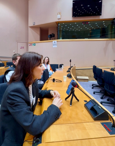 Woman in a blazer speaks into a small microphone on a wooden conference table. People are seated behind her, with a large screen in the background.